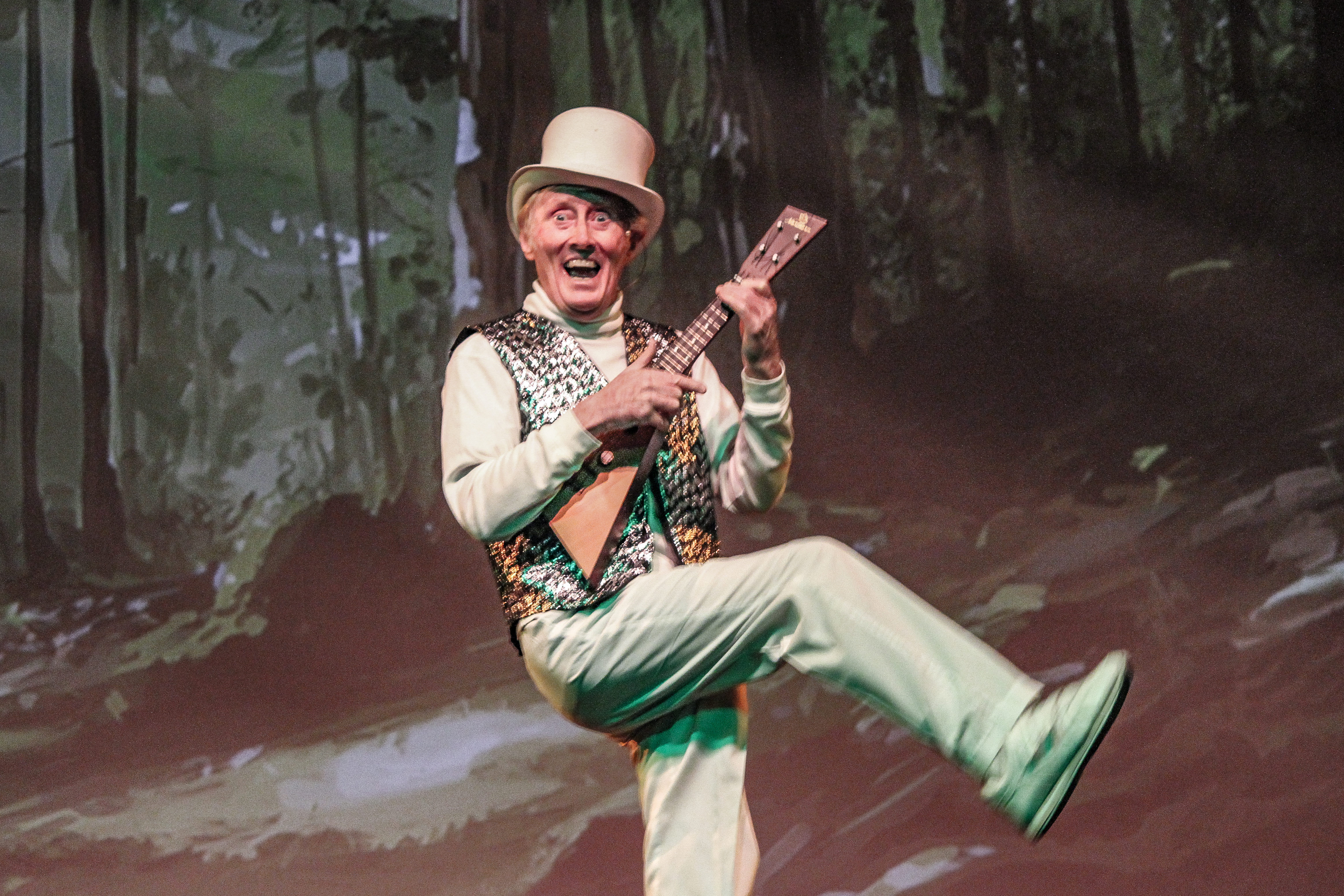 Robin Jarman playing a small stringed instrument on stage. His mouth is wide open and he is wearing a top hat with a sparkling vest. He is kicking one leg out across his body. 